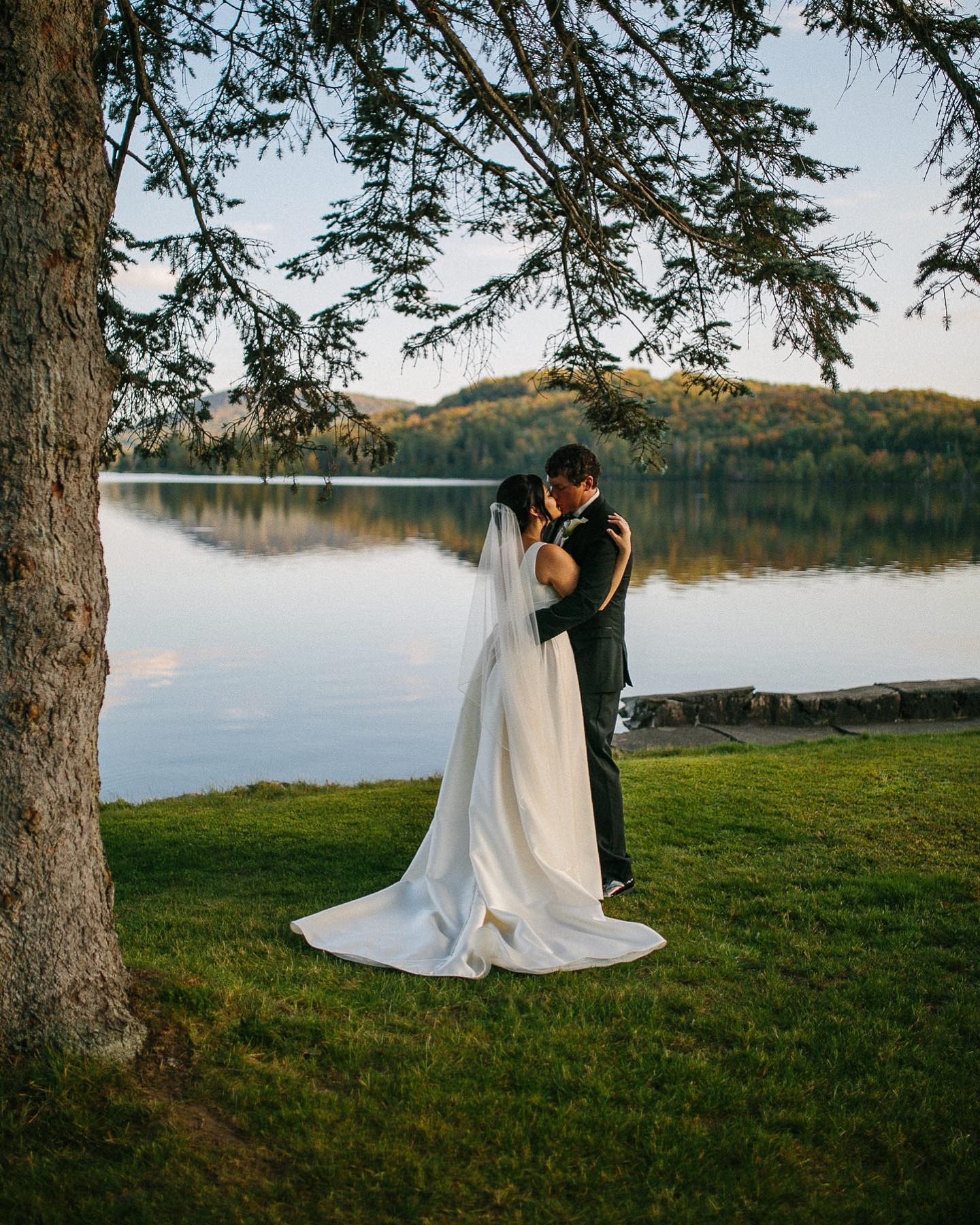 Bride and groom embrace and kiss beside a calm lake, with a large tree in the foreground and forested hills in the distance.