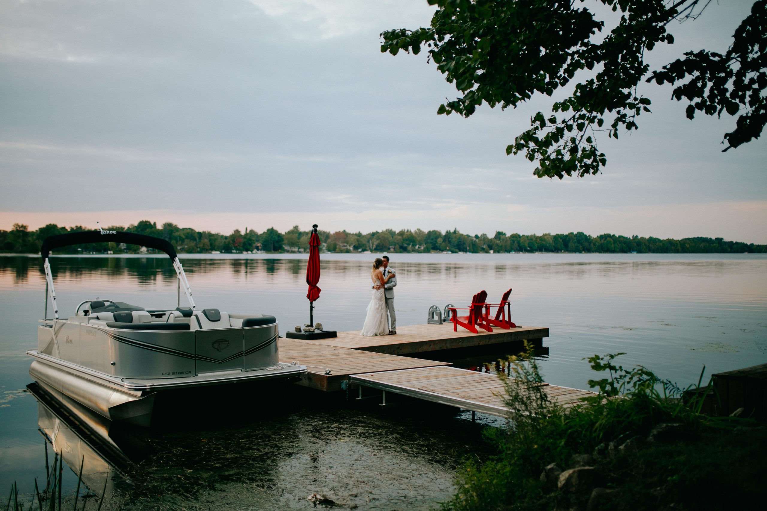 Bride and groom embrace on a wooden lakeside dock, with a pontoon boat nearby and red chairs on the deck behind them.