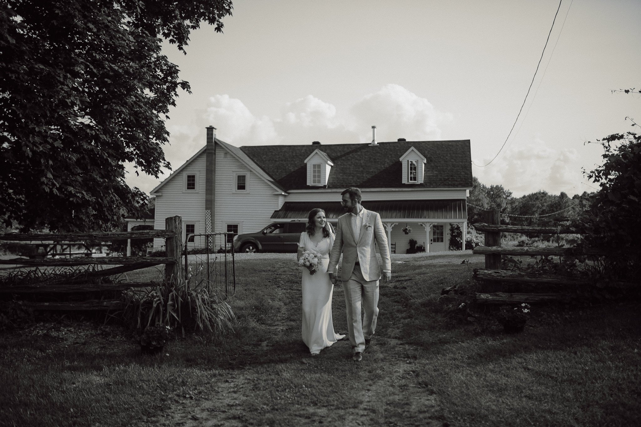 Bride and groom walk hand in hand across a yard in front of a white house, black-and-white wedding photo.