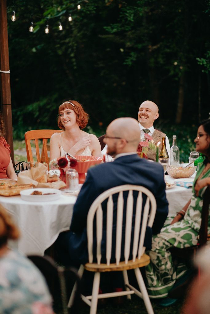 Guests seated at a round outdoor table during a celebration, smiling and enjoying the meal under string lights.
