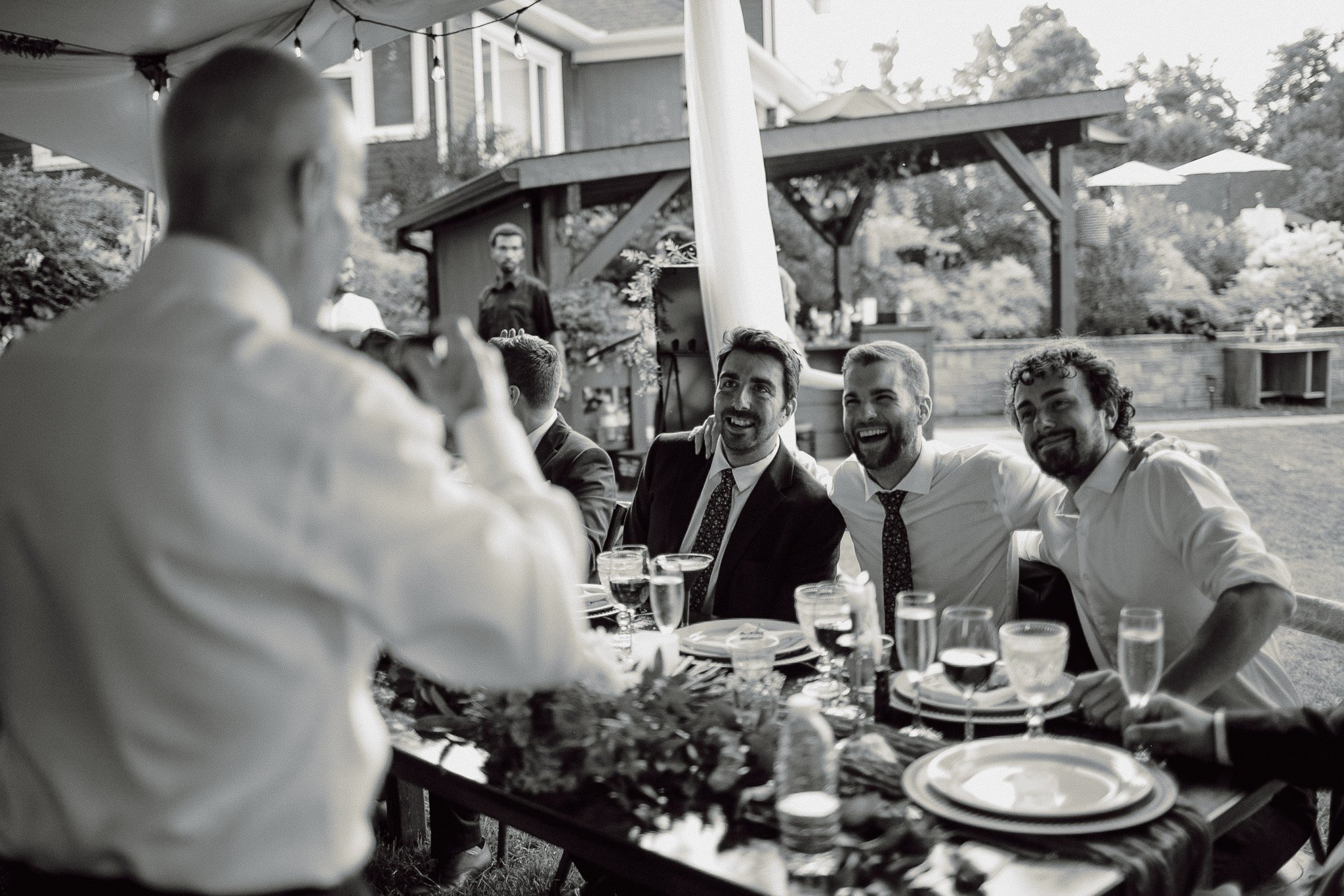 Group of men in suits and ties at an outdoor reception, smiling and toasting drinks around a long table.