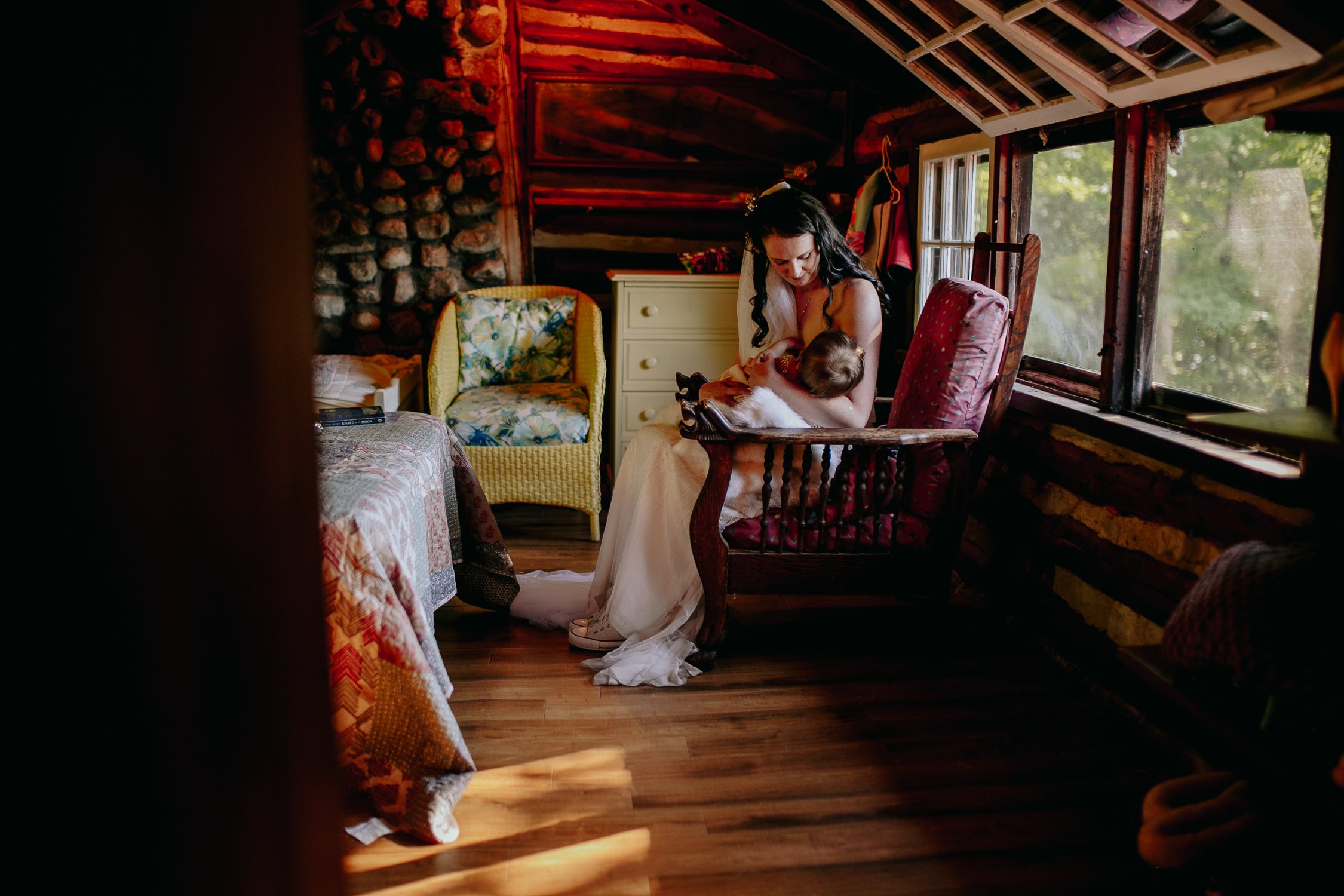 Bride in a white wedding gown cradles a baby in a wooden rocking chair in a cozy rustic attic bedroom bathed in warm light from a window