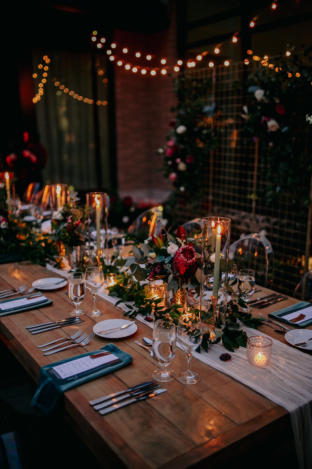 Elegant dinner table set with candlelight, floral centerpiece, and string lights in the background.